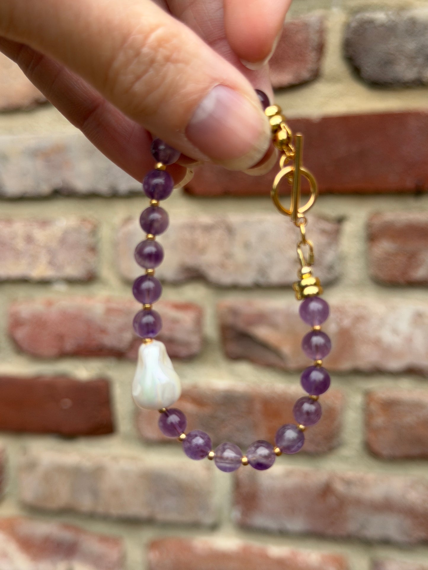Hand holding a beaded bracelet with amethyst and pearl beads and a gold clasp against a brick wall.