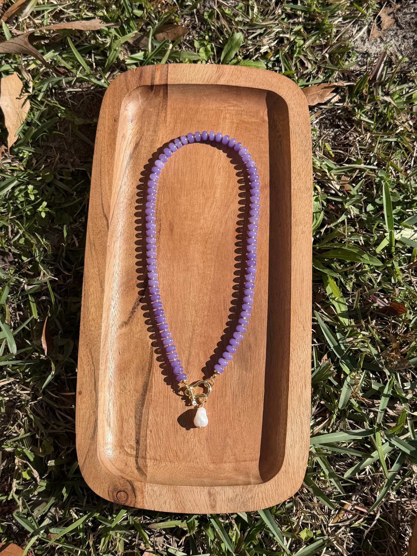 Purple necklace with a pearl charm on a wooden tray outdoors