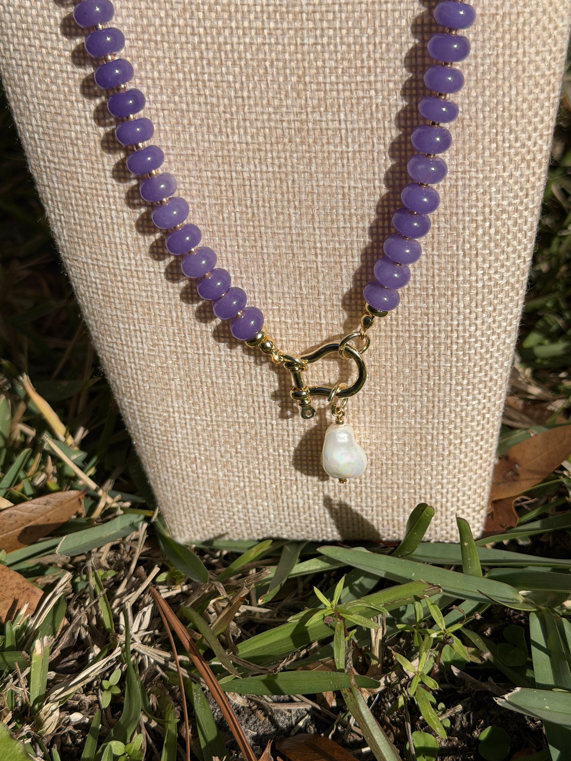 Beaded necklace with purple beads and a pearl pendant on a beige bust, grassy background
