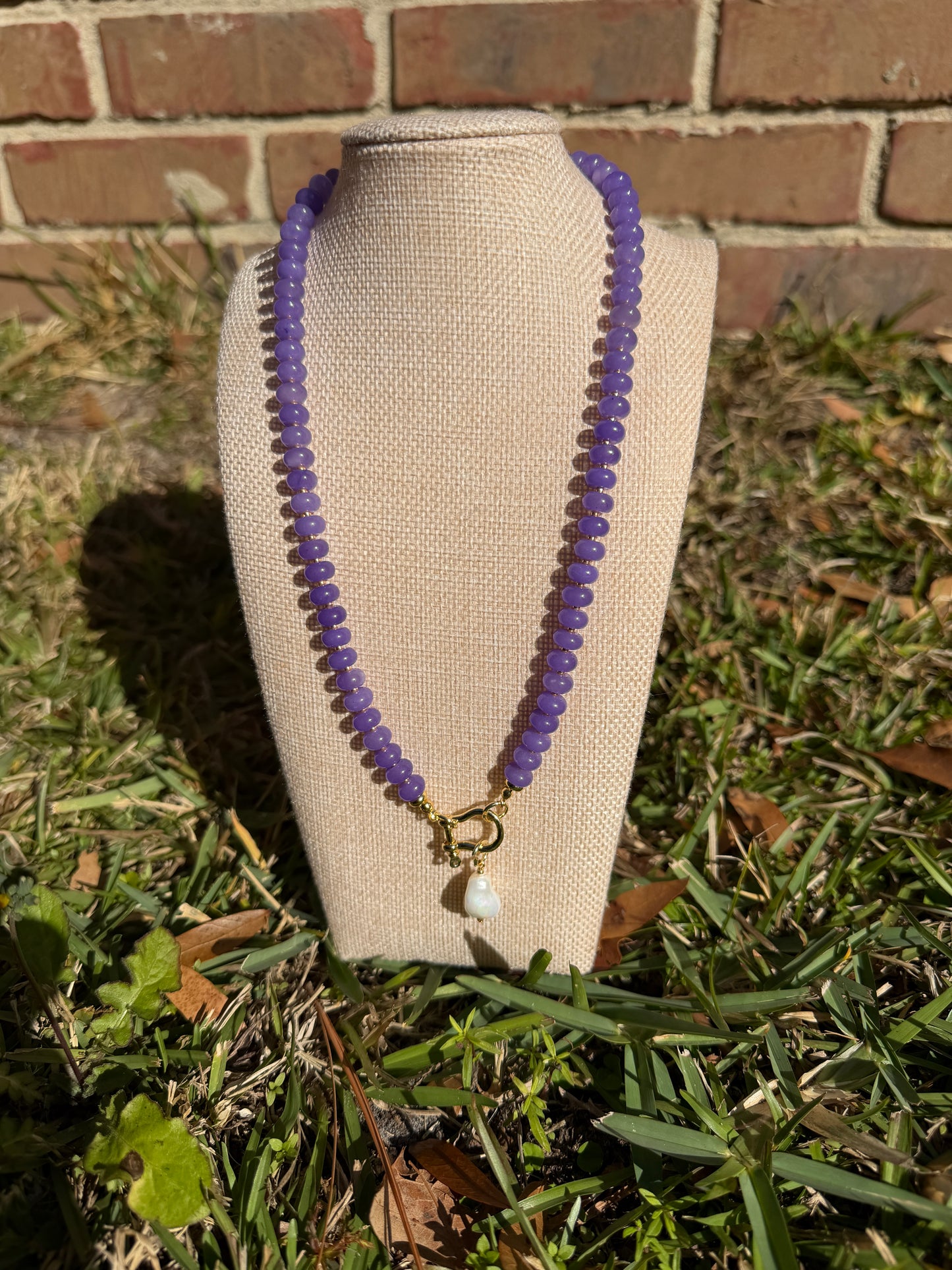 Purple beaded necklace on a beige fabric surface with grass and brick wall in the background